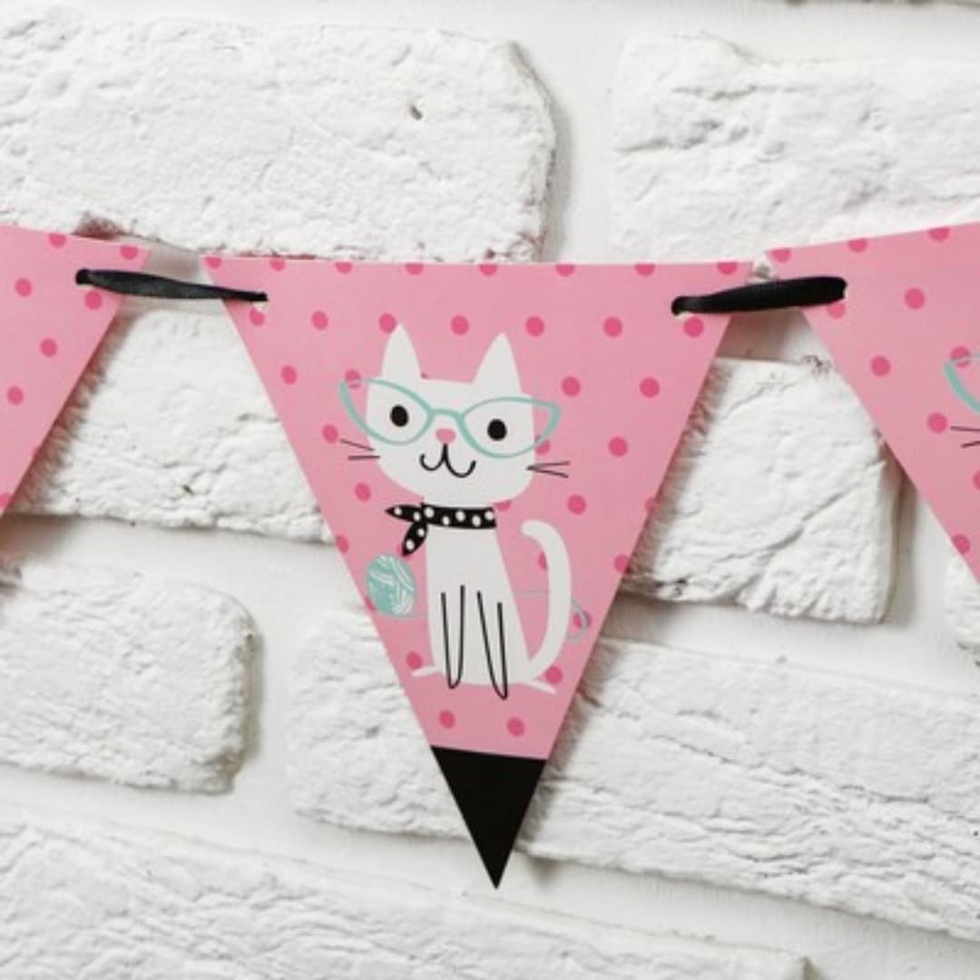 Detalle de banderines de cartulina con diseño de gato blanco sobre fondo rosa pastel