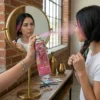 Aplicación de Mechas Rosas Temporales Una mujer joven frente a un espejo circular aplicando el spray de color rosa en una sección de su cabello trenzado. Se ve el reflejo de su rostro mientras rocía el producto en un ambiente luminoso con paredes de ladrillo.