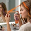 Aplicación de Tinte Temporal Naranja Mujer aplicando spray de color naranja en un mechón de pelo frente al espejo para un resultado vibrante.