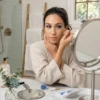 Tutorial de Aplicación de Pestañas Azules Mujer con albornoz beige aplicando una pestaña postiza azul eléctrico con pinzas doradas frente a un espejo de aumento en un baño iluminado.