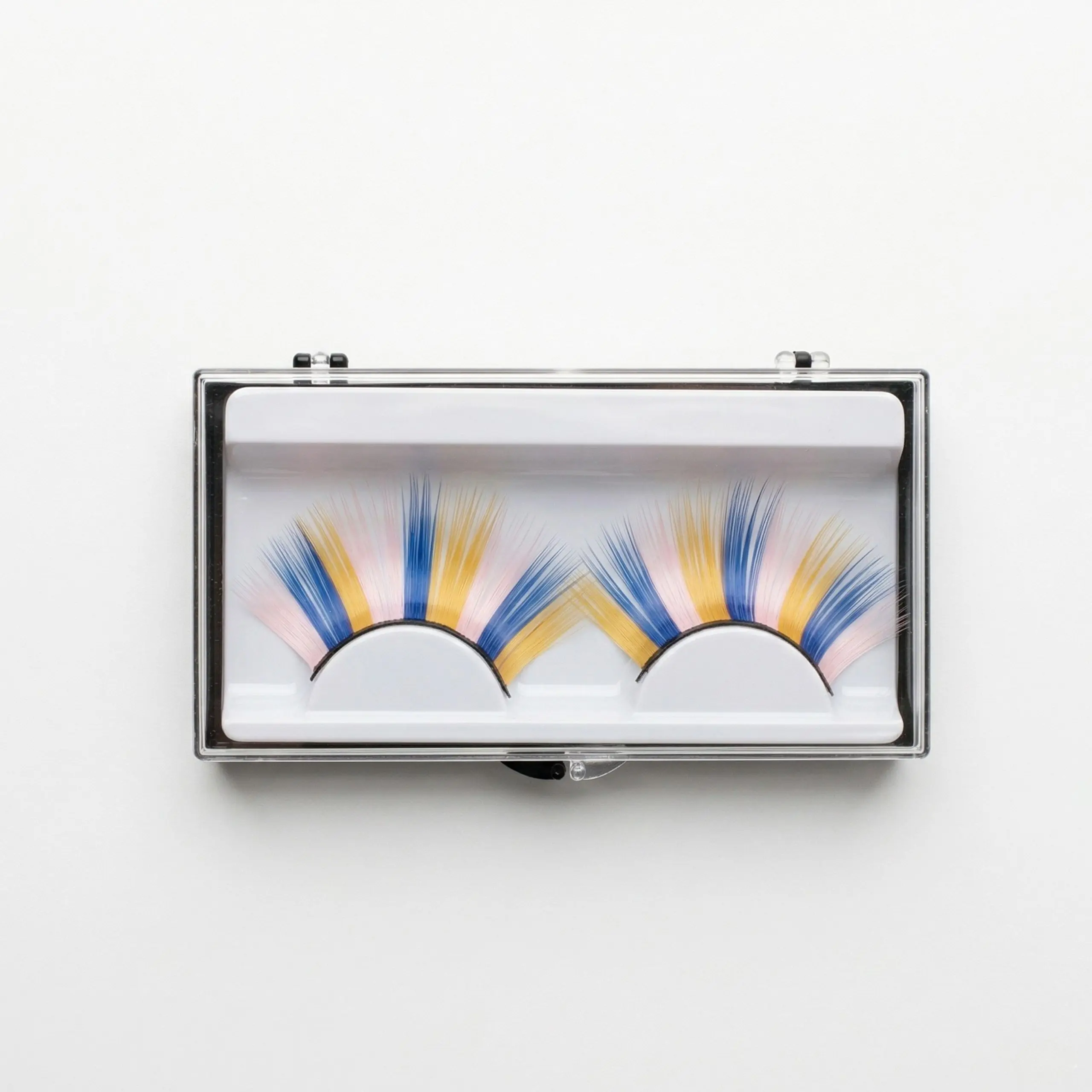 Pack de Pestañas Postizas Multicolor Estuche transparente con un par de pestañas postizas de colores vibrantes en tonos azul, amarillo y rosa pastel sobre fondo blanco.