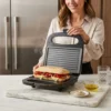 Cómo preparar un panini gourmet con grill de contacto Mujer preparando un sándwich de jamón y pimientos en una prensa de panini eléctrica en una cocina moderna.