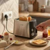 Tostadora Kiwi Acero Inoxidable – Uso Diario Tostadora de acero inoxidable Kiwi con ranuras anchas preparando dos rebanadas de pan tostado con humo saliendo, sobre una mesa de cocina de madera junto a un huevo pasado por agua y mermelada.