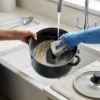 Persona lavando una olla esmaltada negra bajo el grifo con esponja